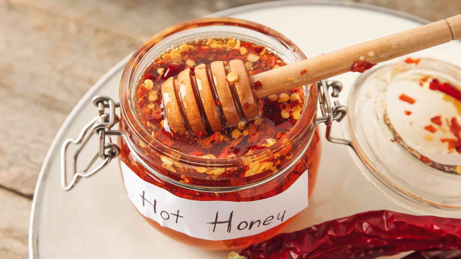 A jar labeled "Hot Honey" filled with honey and chili flakes, with a honey dipper resting on top and a dried chili pepper nearby.