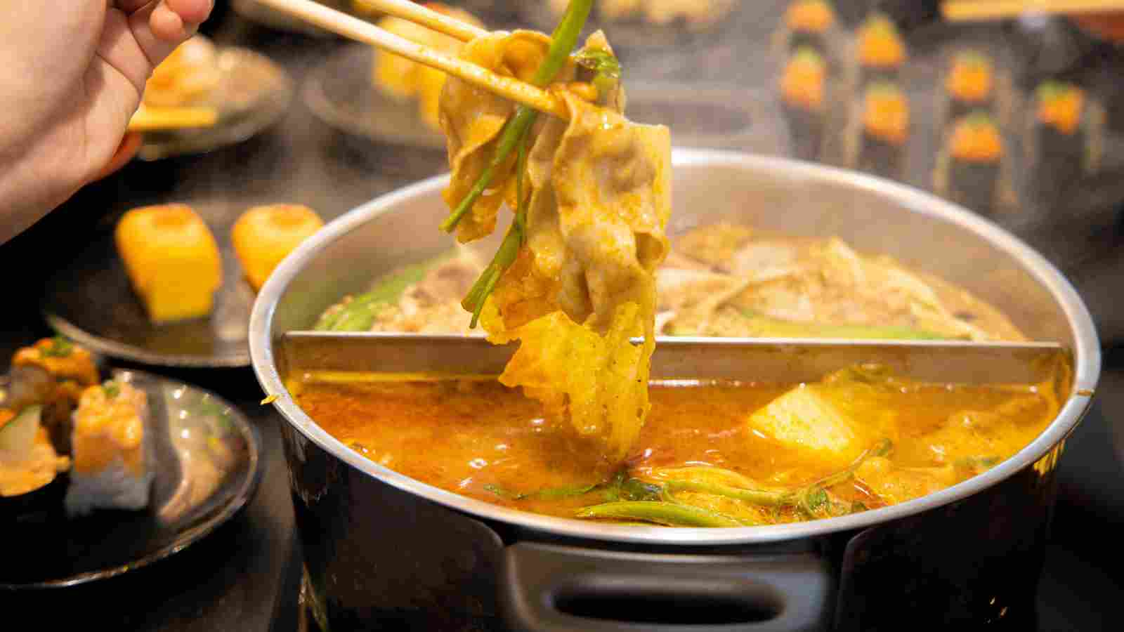 A person uses chopsticks to lift cooked meat and vegetables from a divided hot pot filled with steaming broth, with side dishes visible in the background.