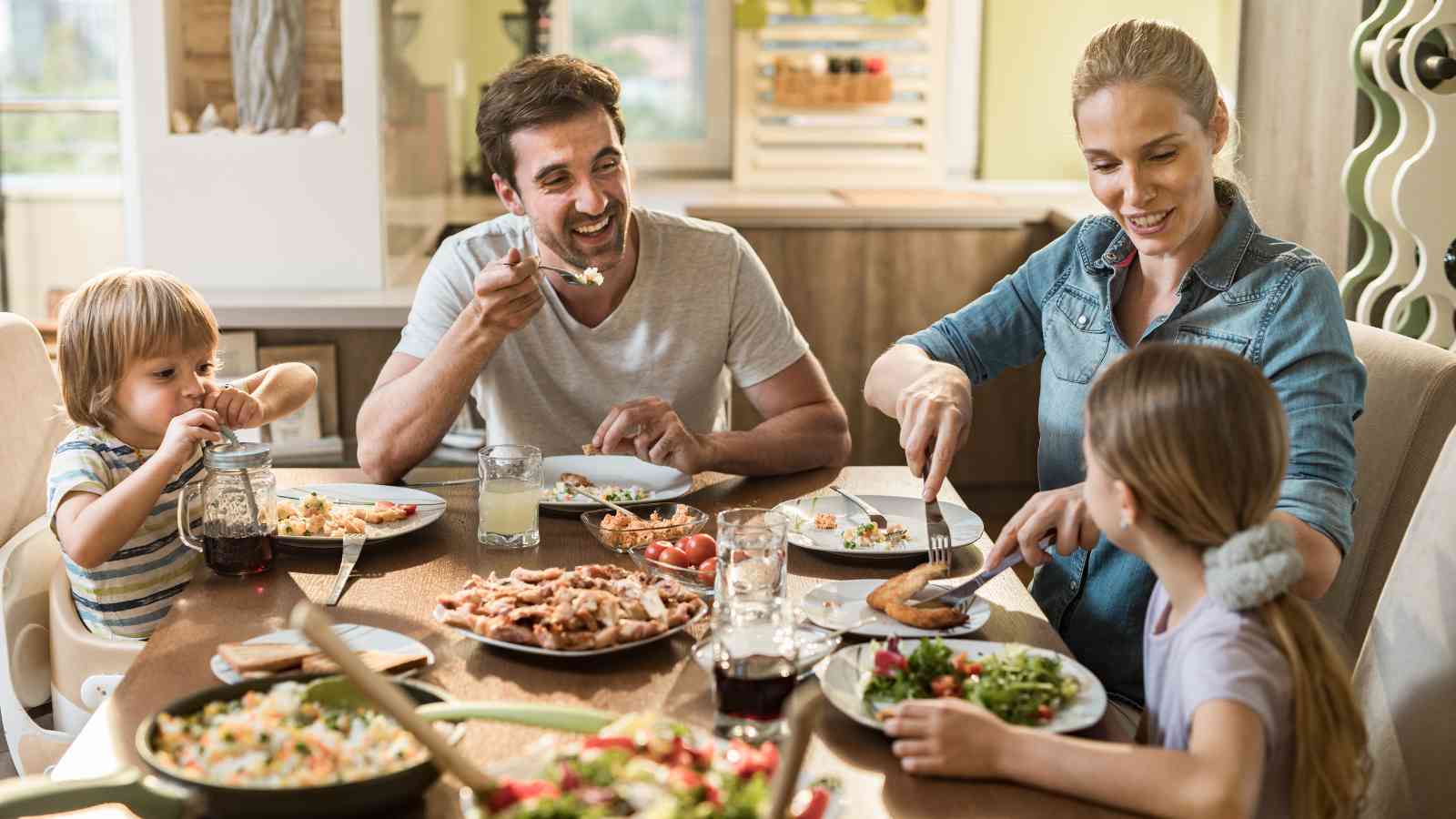 A family of four sits at a dining table, sharing a meal together. The parents serve food to their two children, and plates of salad and meat are spread across the table.