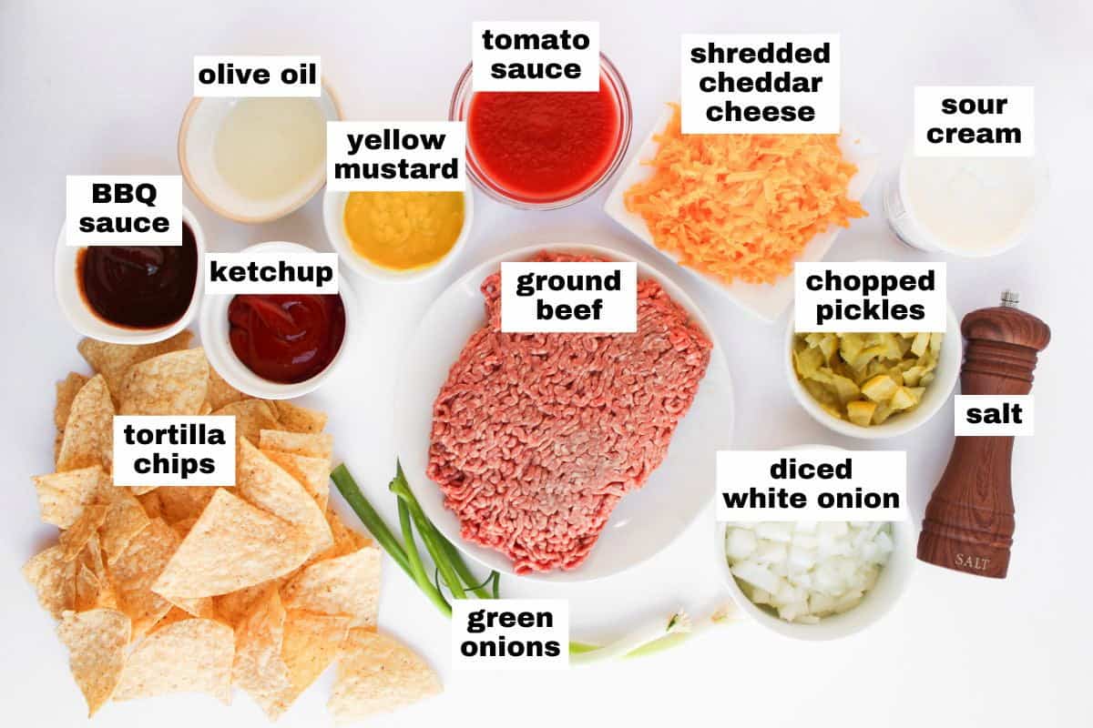 Ingredients arranged on a white surface include ground beef, tomato sauce, shredded cheddar cheese, sour cream, salt, diced onion, pickles, green onions, tortilla chips, ketchup, mustard, and BBQ sauce.