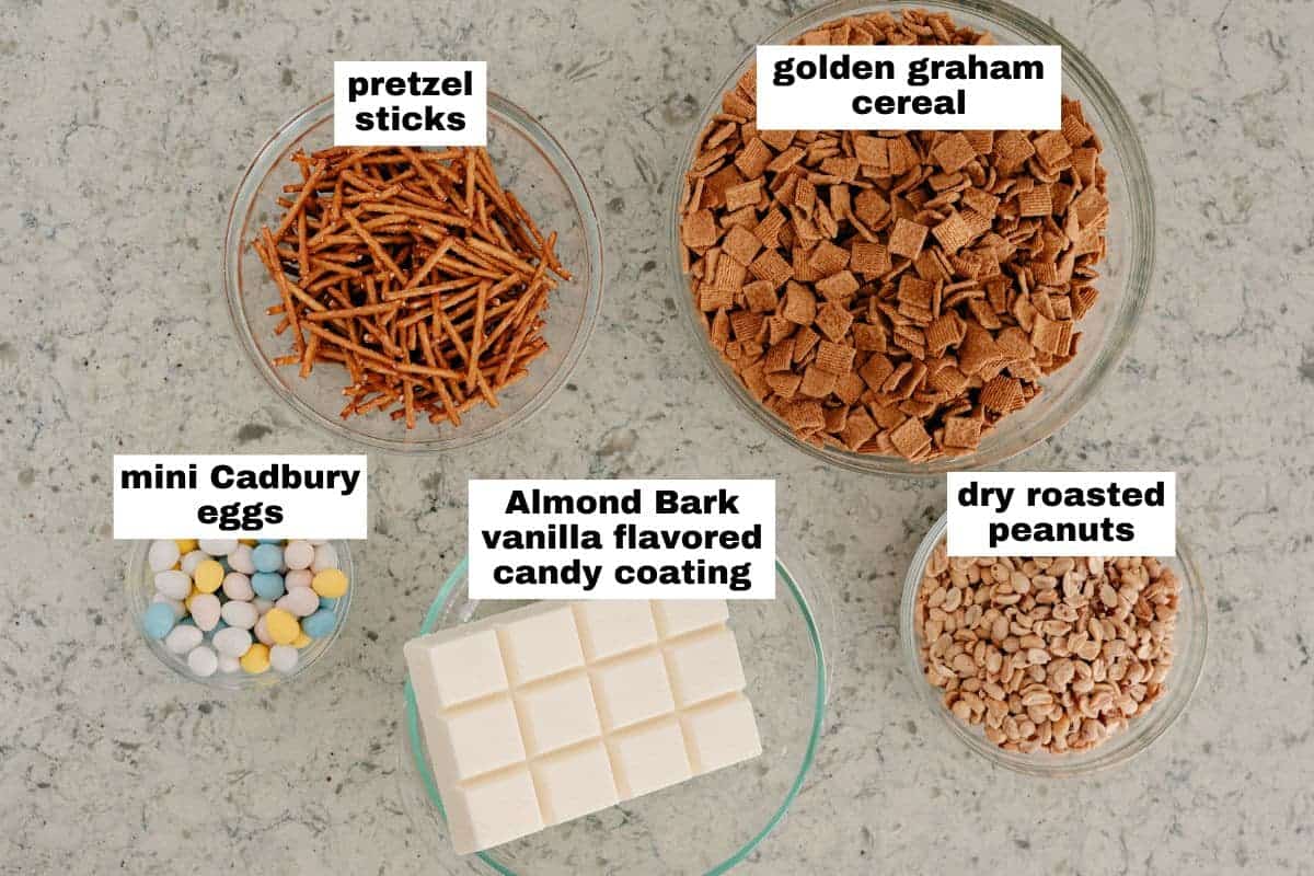 Five bowls display ingredients: pretzel sticks, golden graham cereal, mini Cadbury eggs, Almond Bark vanilla candy coating, and dry roasted peanuts on a countertop.