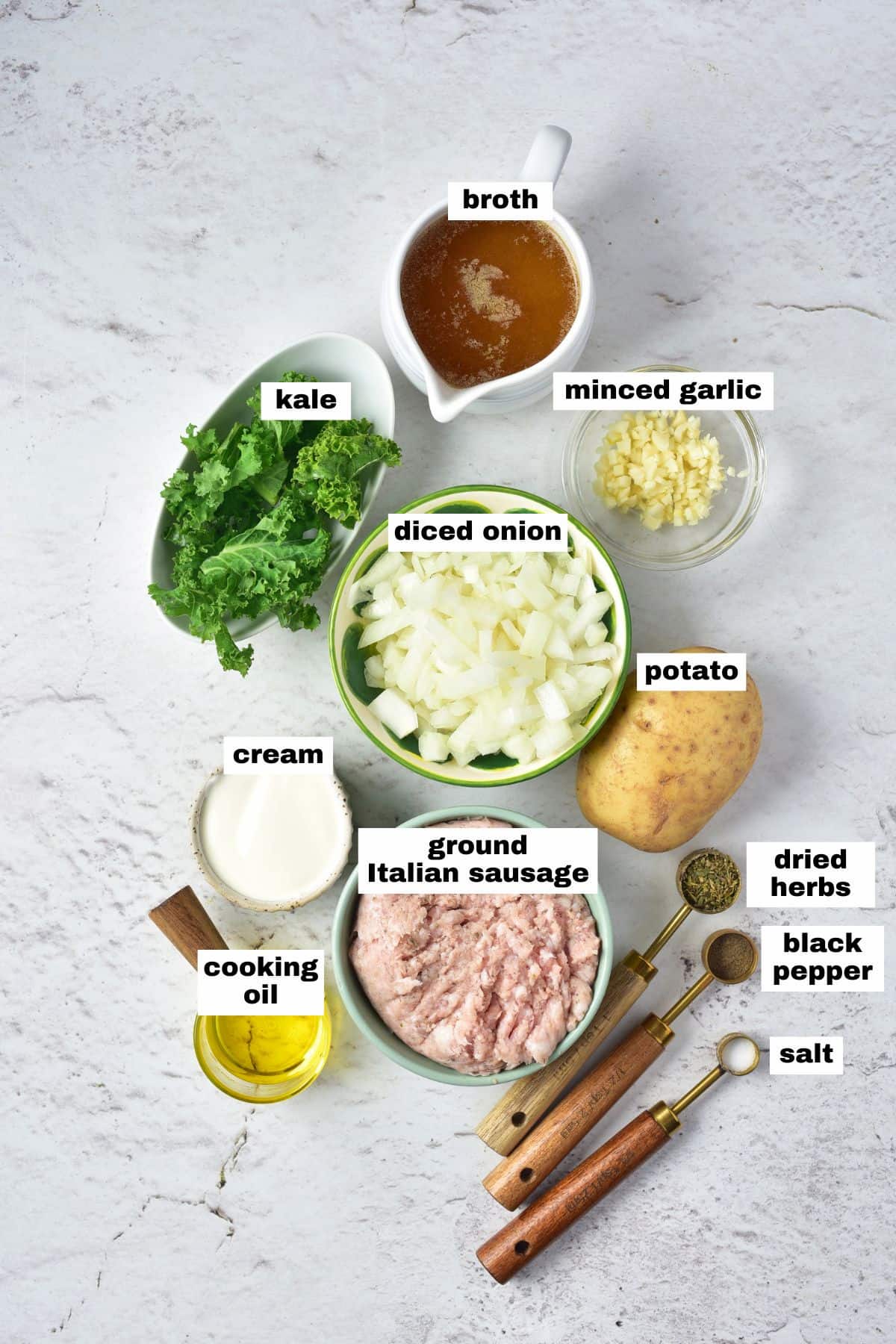 Top-down view of ingredients on a white surface: broth, minced garlic, diced onion, kale, potato, cream, ground Italian sausage, cooking oil, dried herbs, black pepper, and salt.