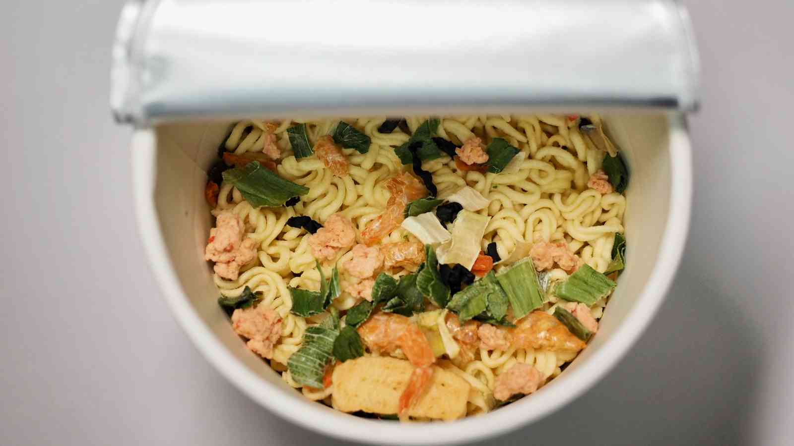Opened instant noodle cup showing dry noodles, dehydrated vegetables, and seasoning inside, viewed from above.
