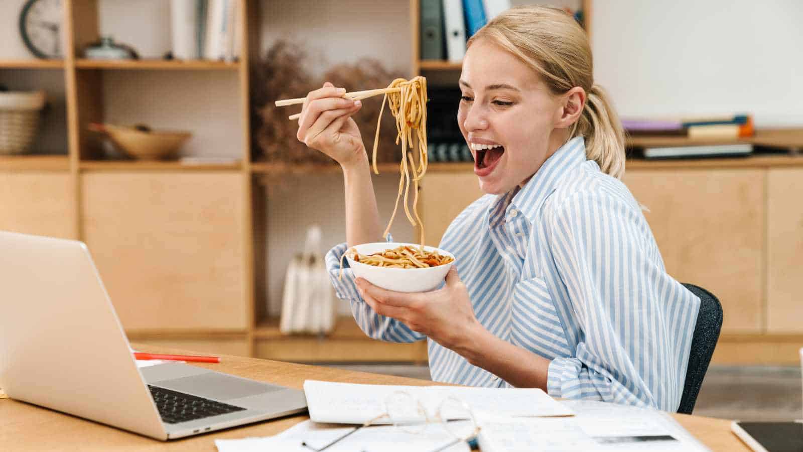 Woman sitting at a desk eating noodles with chopsticks while working on a laptop, with papers and glasses on the desk.
