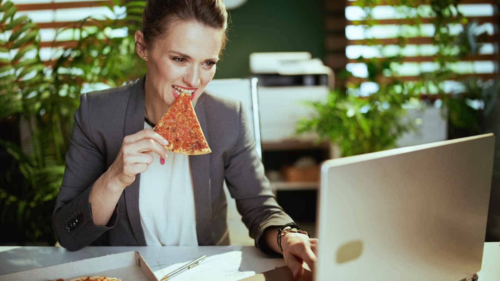 A woman in business attire eats pizza while looking at a laptop at her desk, with plants and paperwork visible in the background.