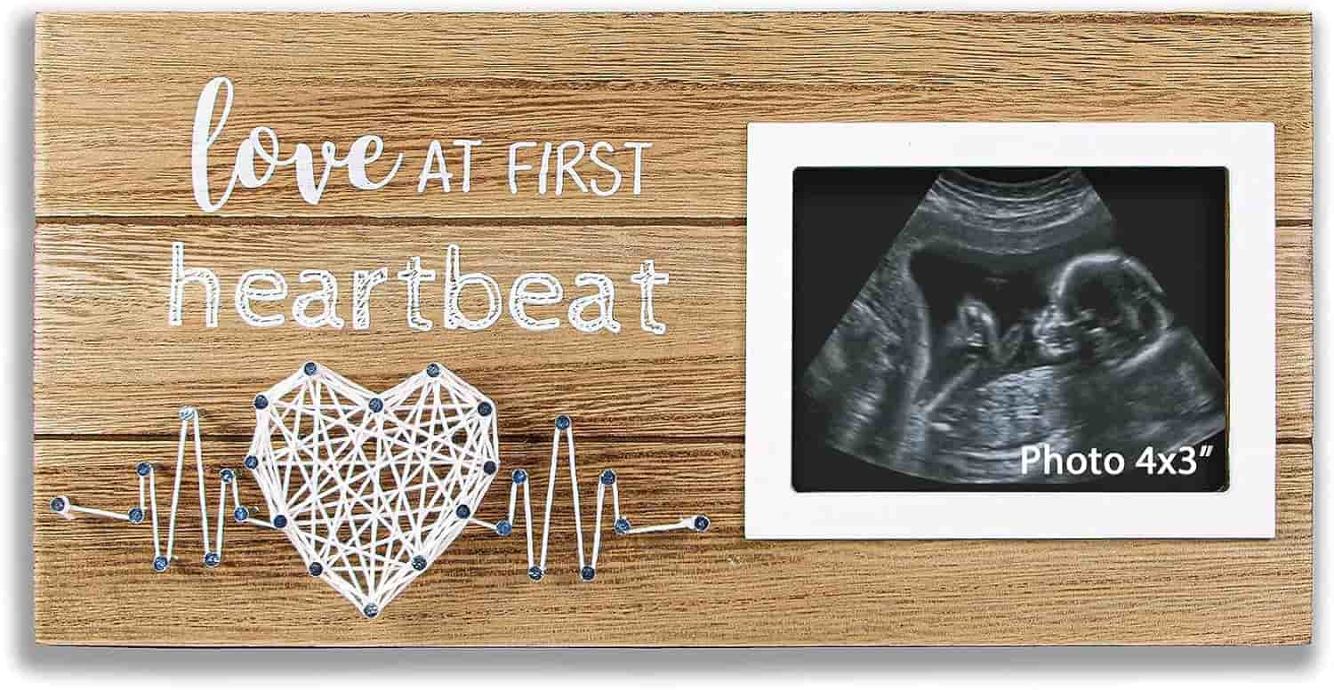 A wooden photo frame with the words "love at first heartbeat," featuring a string art heart and a 4x3 inch ultrasound photo of a fetus.