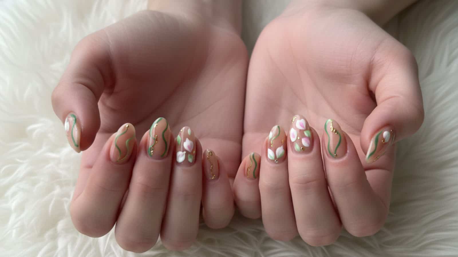 Two hands with long fingernails featuring a pastel nail art design of wavy lines, small gold dots, and white flowers, displayed on a soft white surface.