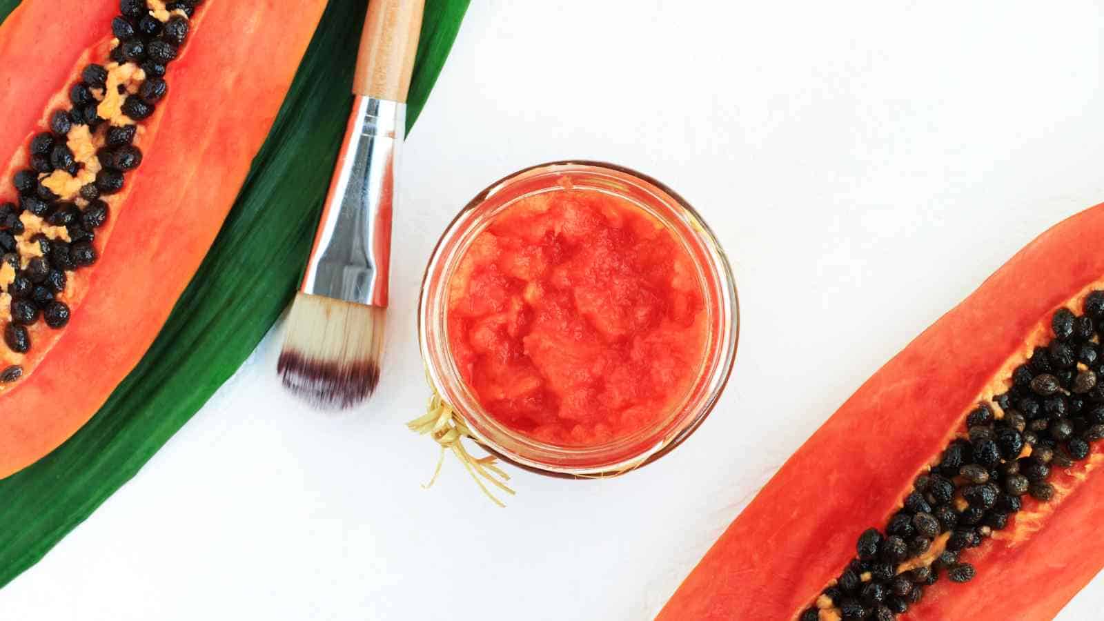 A jar of papaya pulp is placed between two halved papayas with seeds exposed, a green leaf, and a facial brush on a white background.