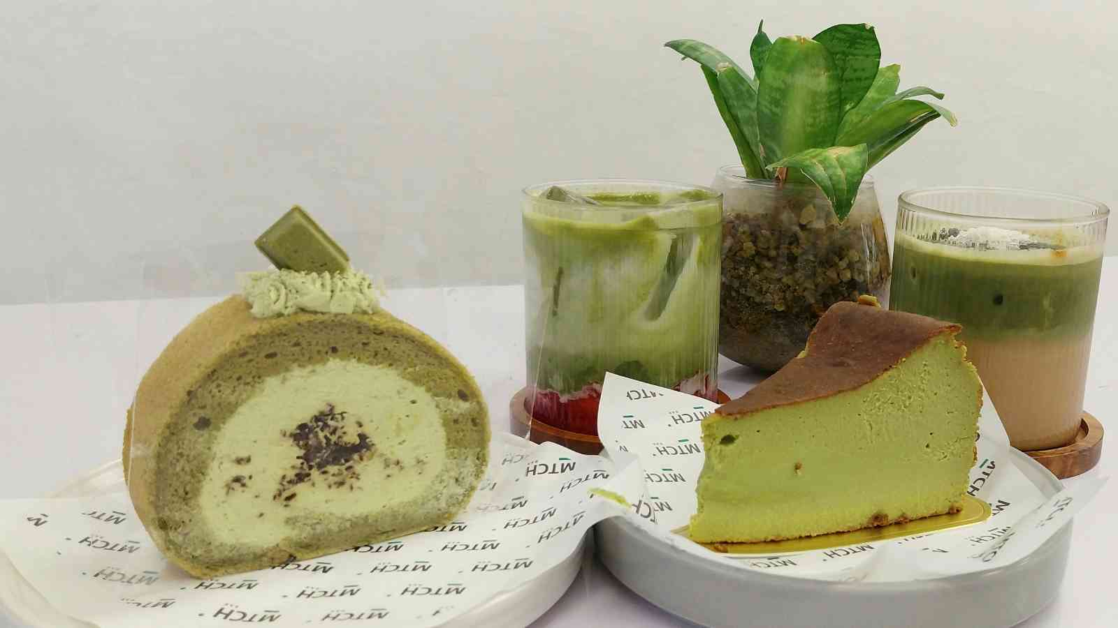 A matcha roll cake, matcha cheesecake, two matcha drinks, a small potted plant, and a strawberry dessert are arranged on a table with branded paper.