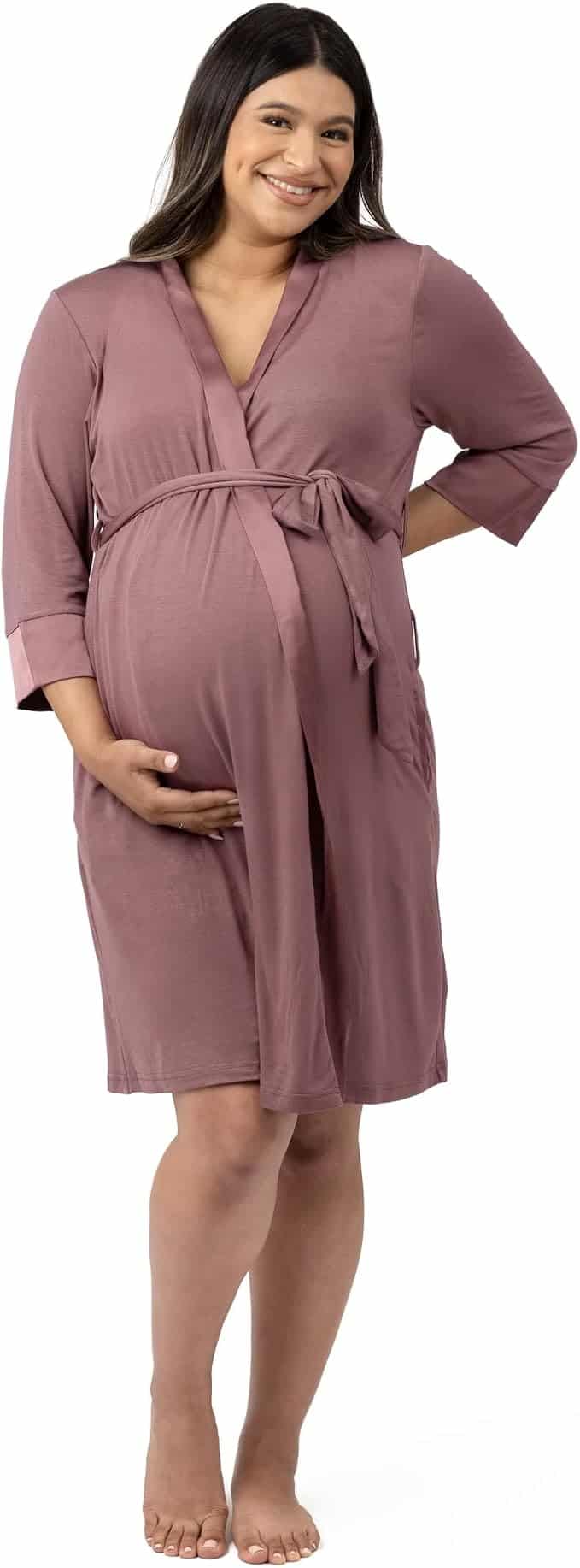 Pregnant woman wearing a mauve knee-length robe with a tie belt, smiling and holding her belly, standing barefoot against a white background.