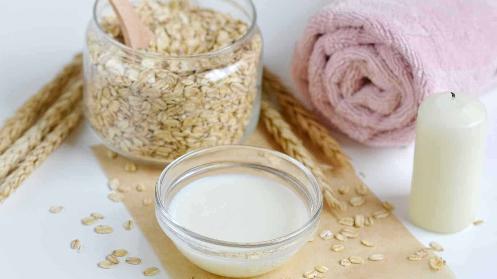 A bowl of milk, a jar of oats with a wooden spoon, a rolled pink towel, a white candle, and oat grains on a white surface.