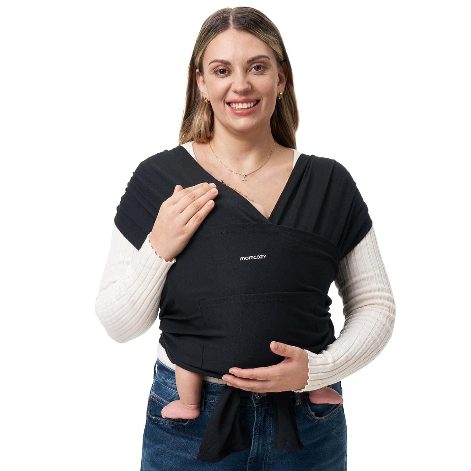 A woman smiles at the camera while wearing a black baby carrier wrap with a baby inside; only the baby's legs and feet are visible.