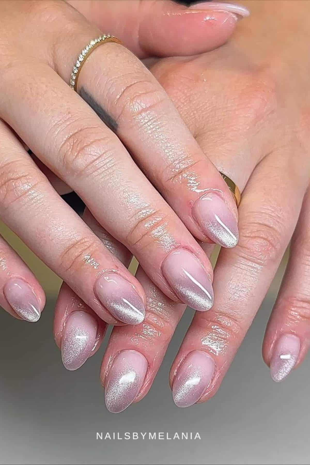 Hands with almond-shaped nails featuring a shimmery pale pink polish. Two gold rings are visible on the fingers.