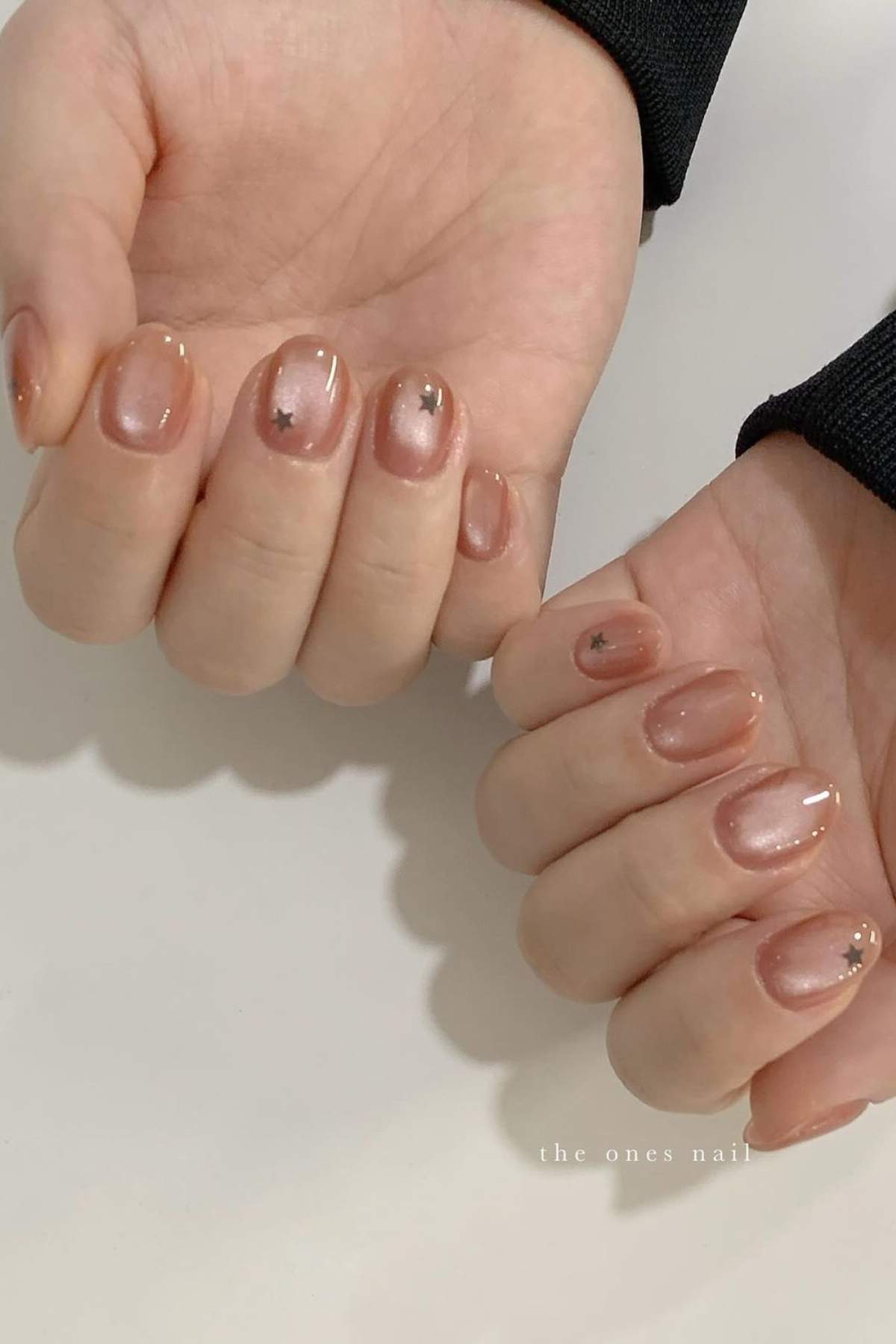 Close-up of hands with short, rounded nude nails featuring a glossy finish and small black star-shaped accents on a few nails.