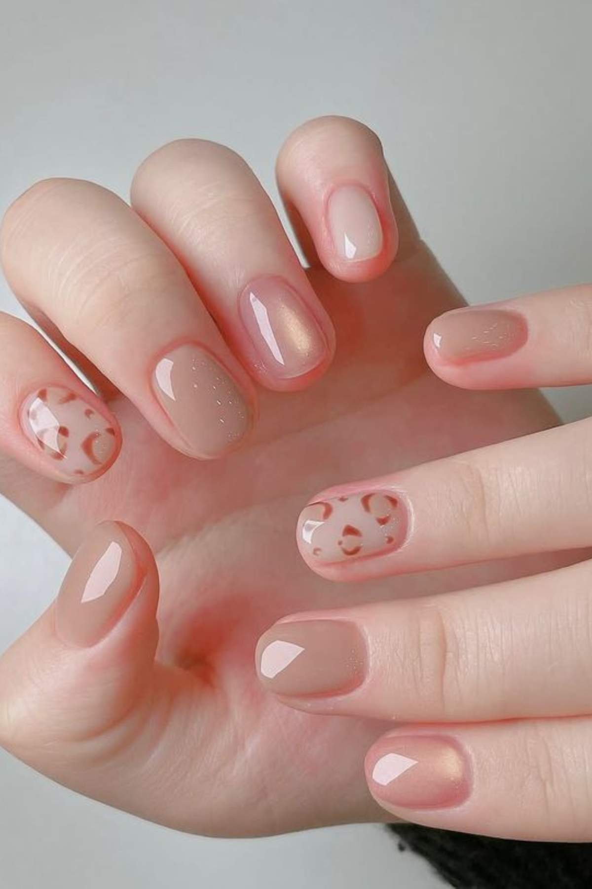 Close-up of hands with short, rounded nails painted in nude and light pink polish, with accent nails featuring a light brown abstract pattern.