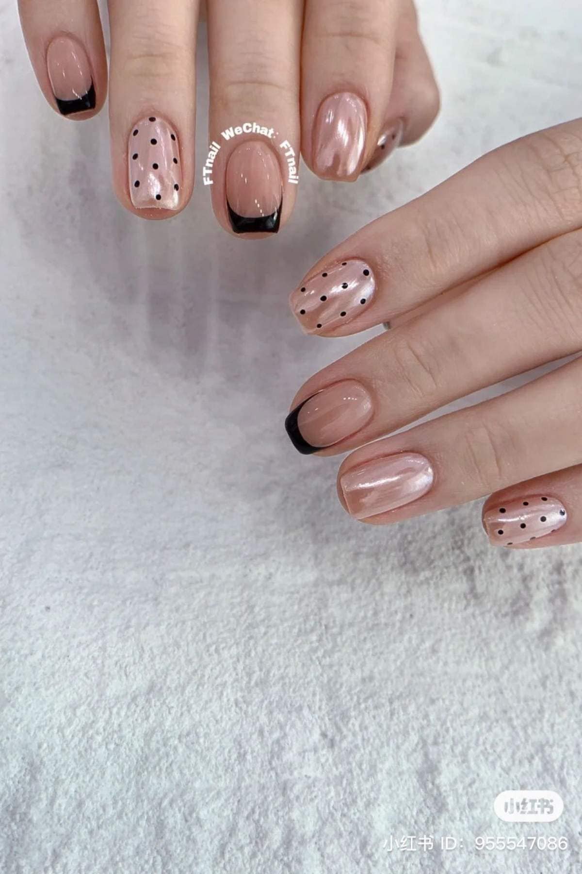 Hands with short, nude-colored nails featuring black French tips and black polka dot designs on some nails, displayed on a light surface.
