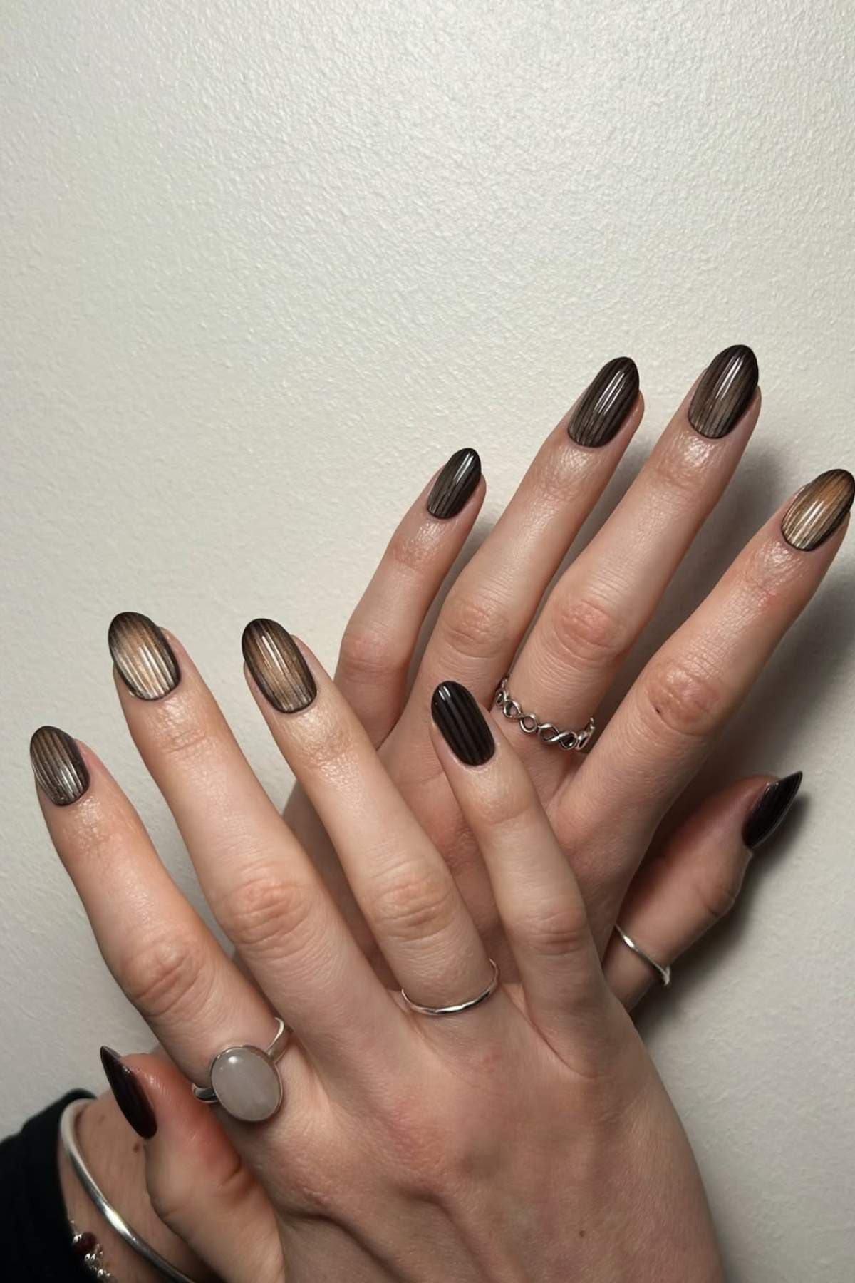 Hands with almond-shaped nails featuring a brown and beige ombre design with vertical lines, wearing several silver rings, against a plain light background.