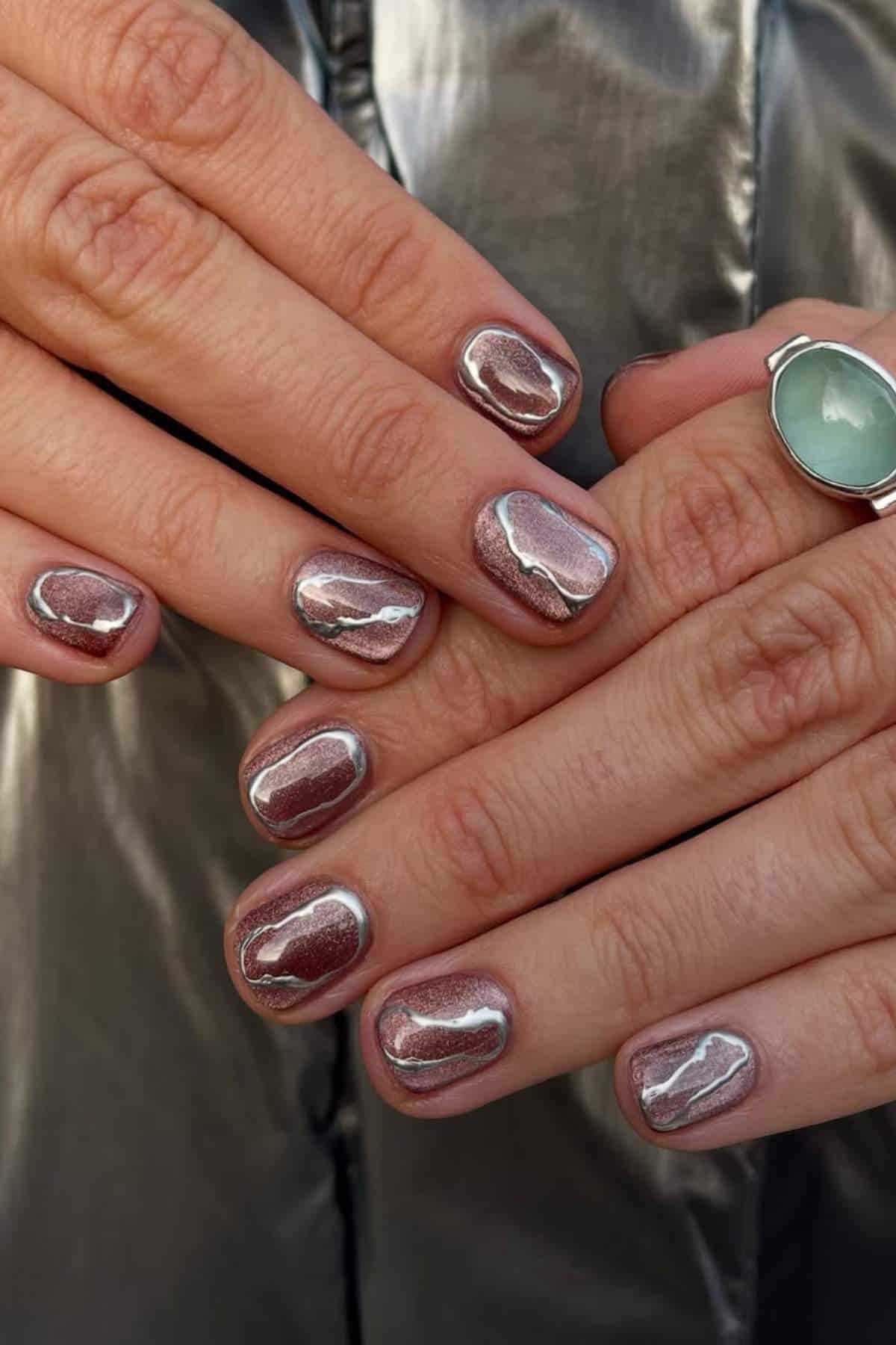 Close-up of hands with short, oval nails painted in a reflective chrome metallic polish; a silver ring with a green stone is on one finger.