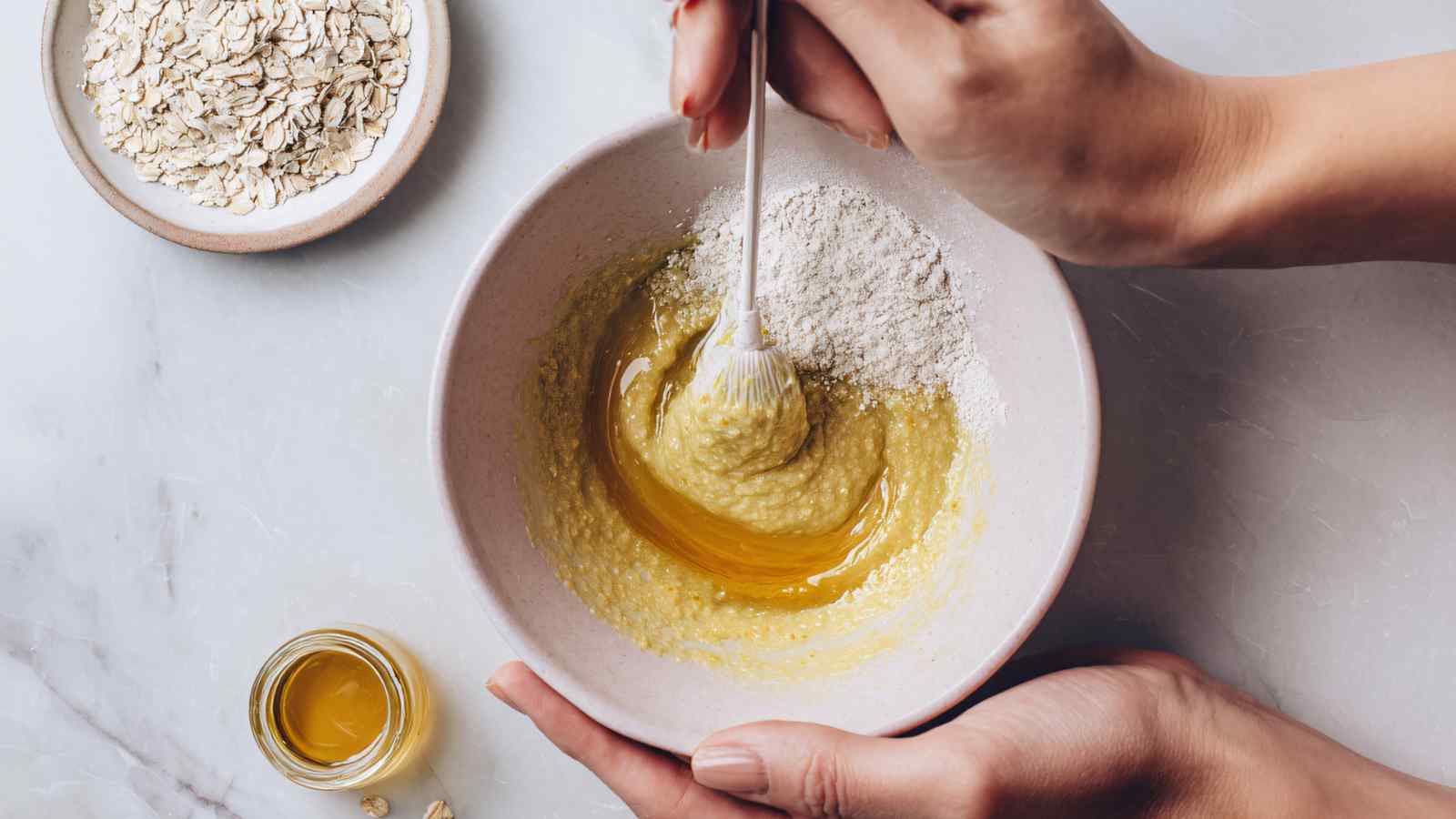 Hands mix flour into a yellow batter in a bowl, with a small dish of oats and a jar of oil on a light countertop.