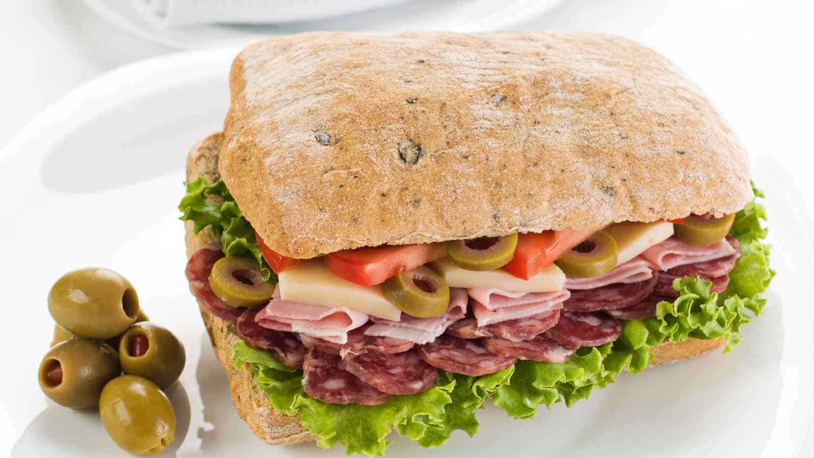 A ciabatta sandwich with lettuce, salami, ham, cheese, tomato, and green olives, served on a white plate with extra green olives on the side.