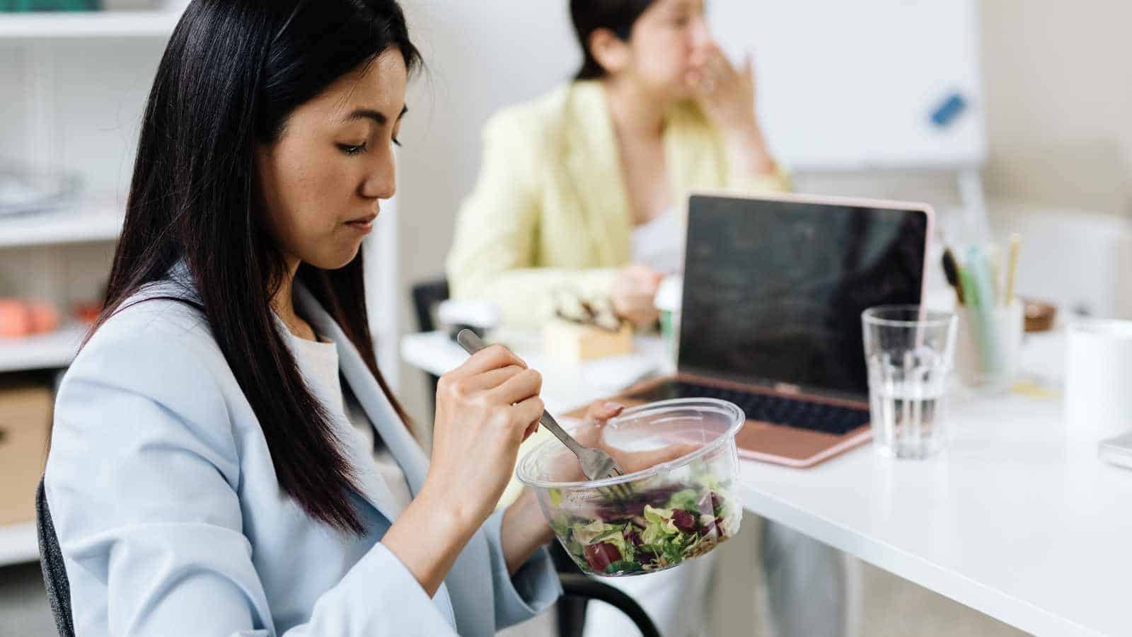 A woman sitting at a desk eating salad from a plastic container, with a laptop, glass of water, and another person working in the background.