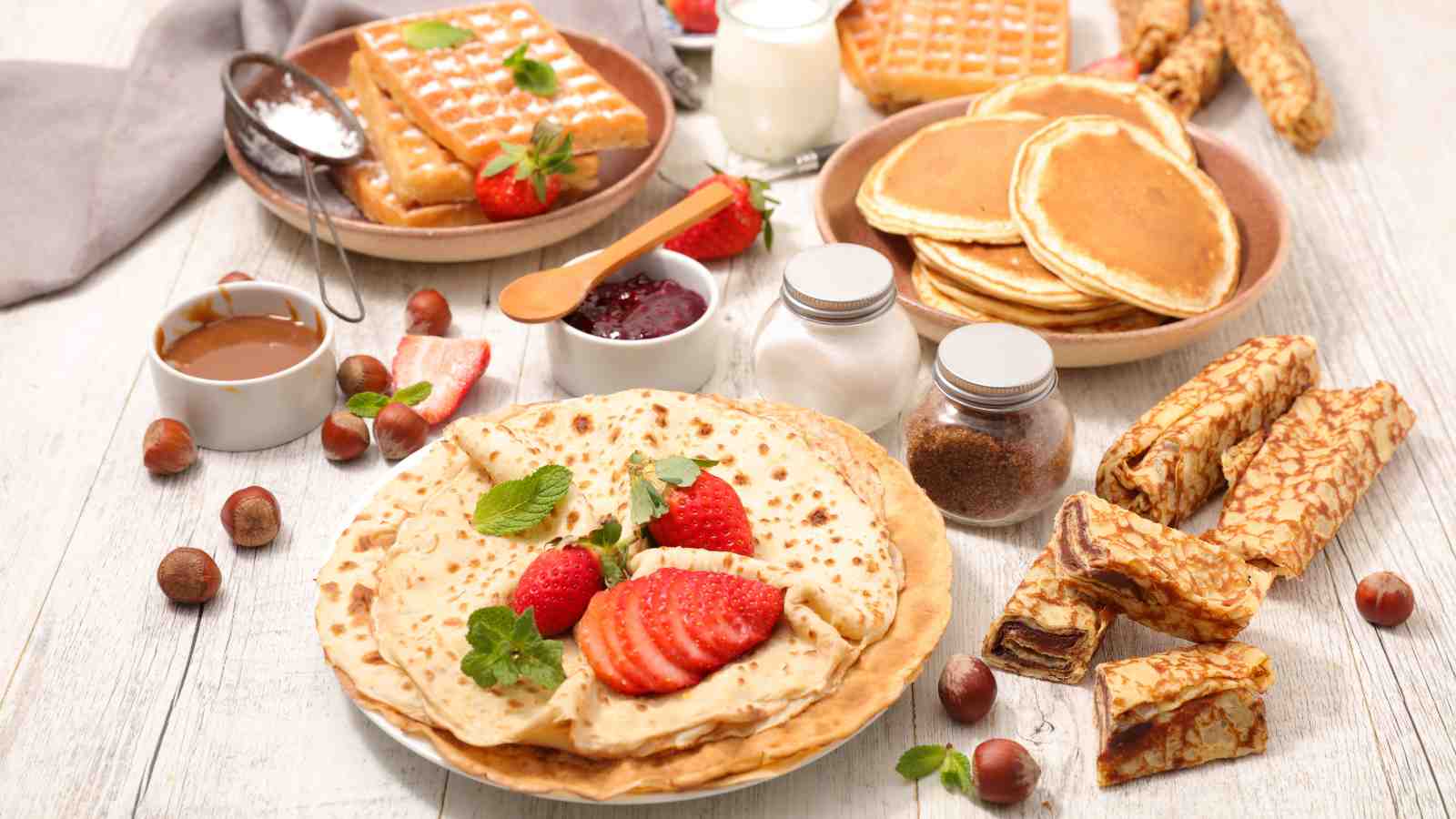 A breakfast spread with crepes, pancakes, waffles, rolled pastries, strawberries, hazelnuts, jam, syrup, milk, and condiments on a white wooden table.