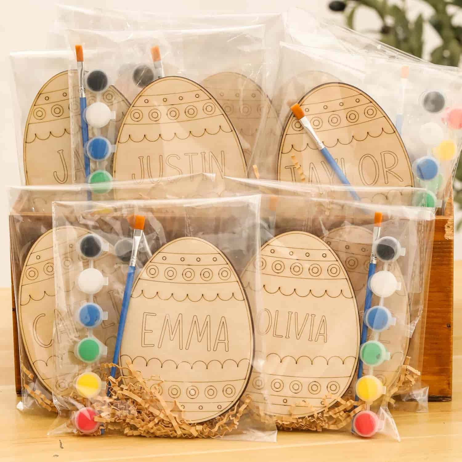 Clear plastic bags contain wooden egg ornaments labeled with names, small paintbrushes, and paint pots, arranged upright in a wooden box on a table.