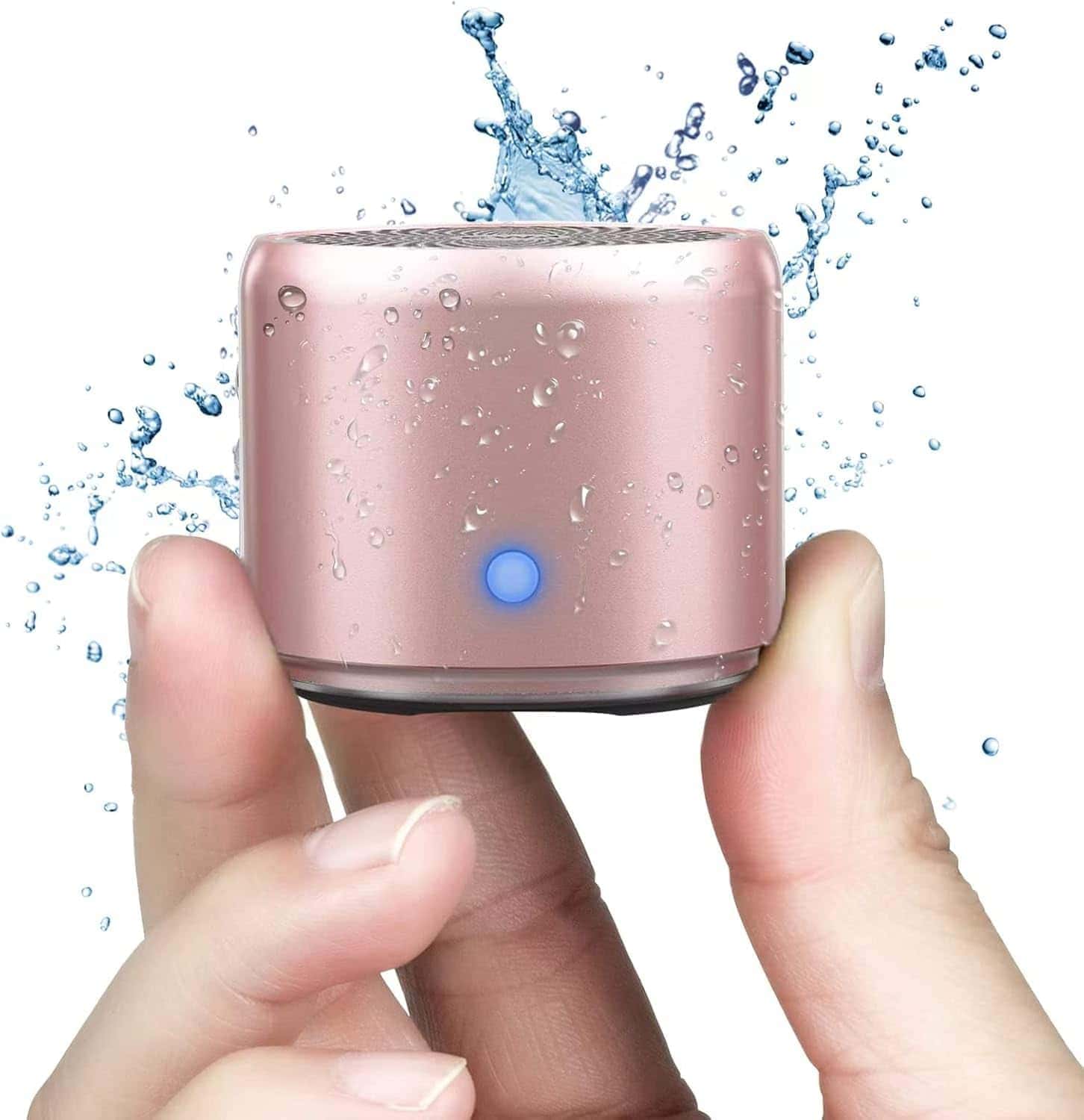 A hand holds a small pink wireless speaker with a blue power light; water splashes behind it, suggesting it is water-resistant.