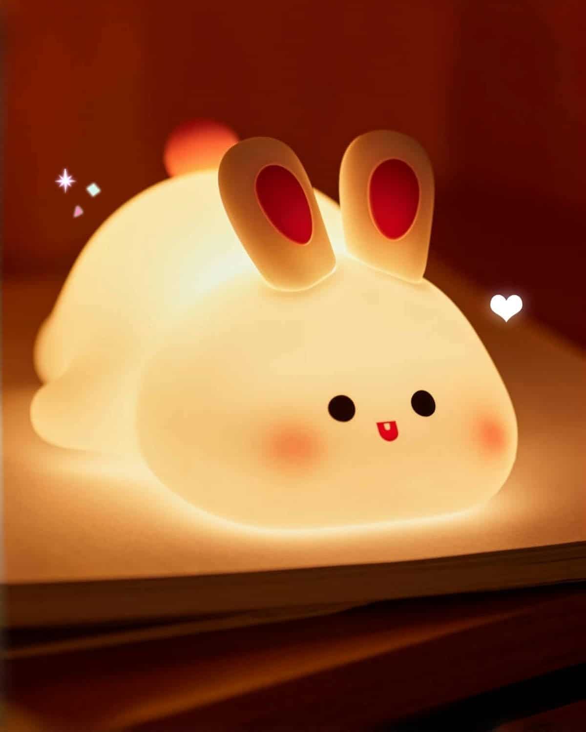 A soft, glowing bunny-shaped night light with rounded features, red-lined ears, and a smiling face sits on a flat surface in a dimly lit room.
