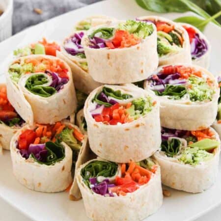A white plate with neatly stacked veggie pinwheel wraps filled with colorful vegetables, next to a small bowl of seeds and scattered spinach leaves.