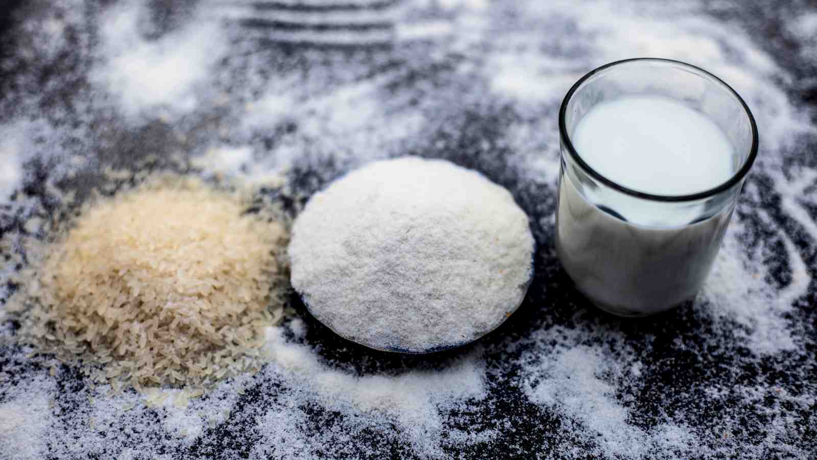 A pile of uncooked rice, a bowl of rice flour, and a glass of milk are arranged in a row on a dark surface dusted with flour.