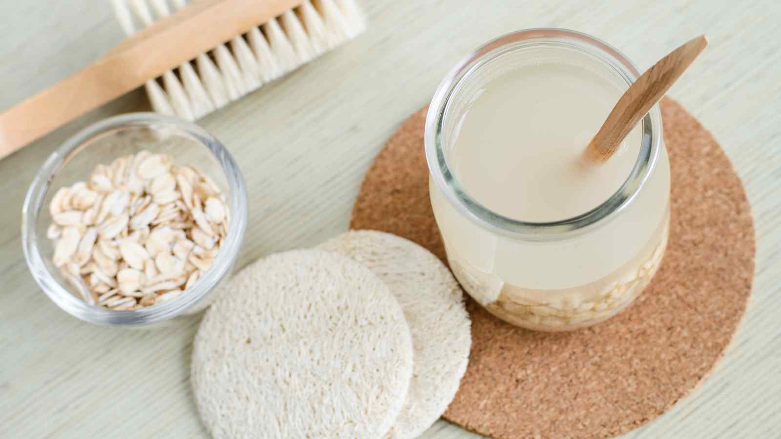 A jar containing a milky liquid with oats and a wooden spoon, placed on a cork mat, next to round cotton pads, a bowl of oats, and a wooden brush.