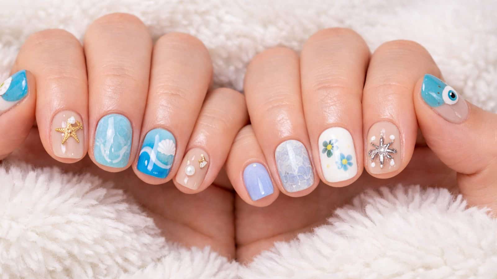 Hands with manicured nails featuring blue, white, and nude designs, including starfish, waves, flowers, gems, and an eye pattern, against a white fuzzy fabric.