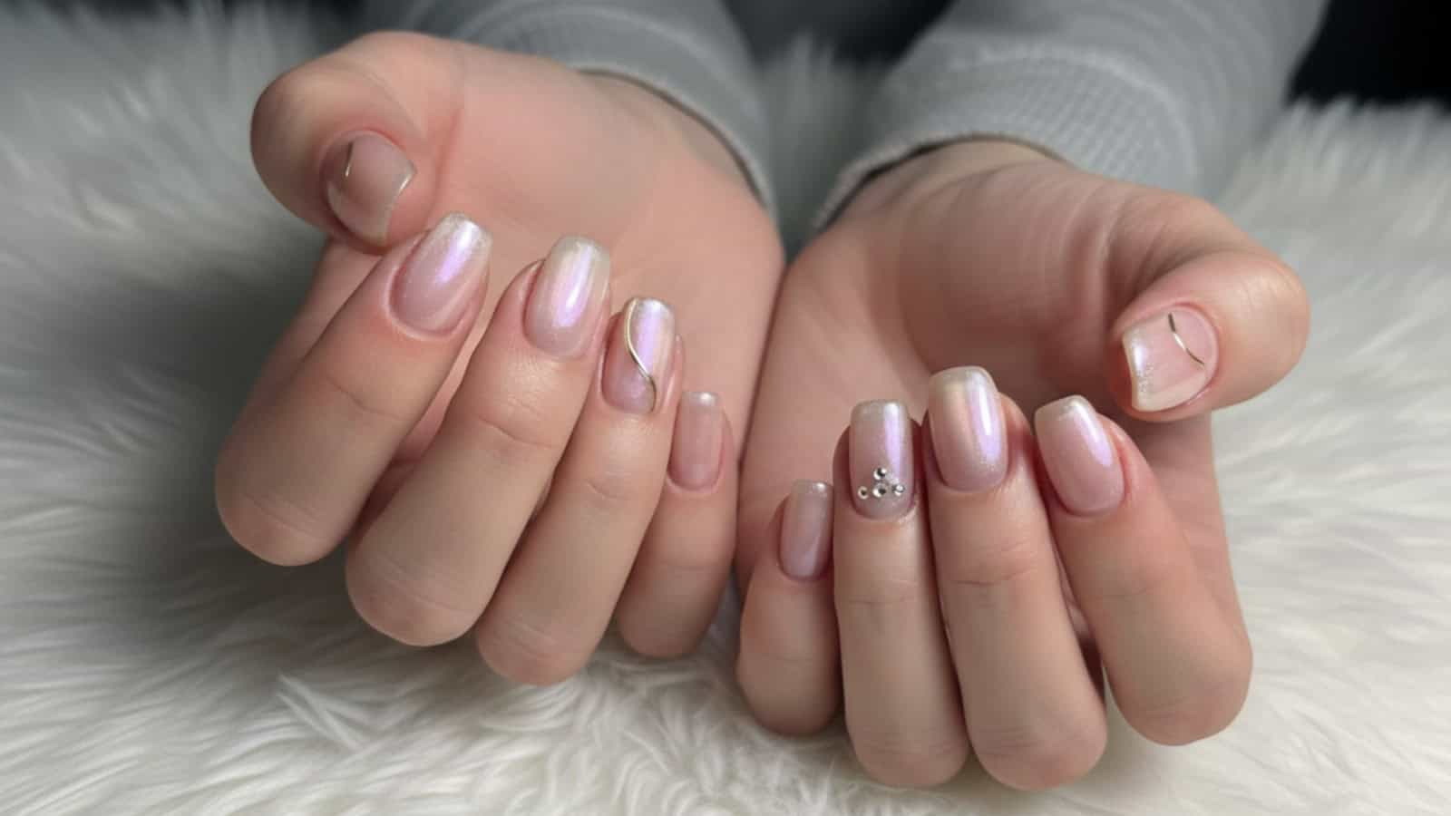 Two hands with short, almond-shaped nails featuring light pink and iridescent polish, minimalist silver lines, and small rhinestone accents, resting on a white fuzzy surface.