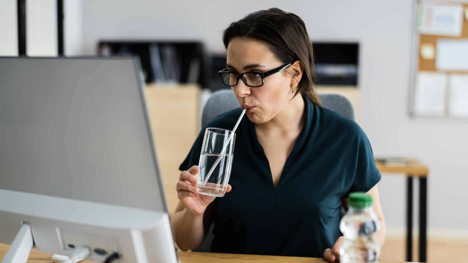A person wearing glasses sits at a desk, drinking water through a straw from a glass while looking at a computer monitor.