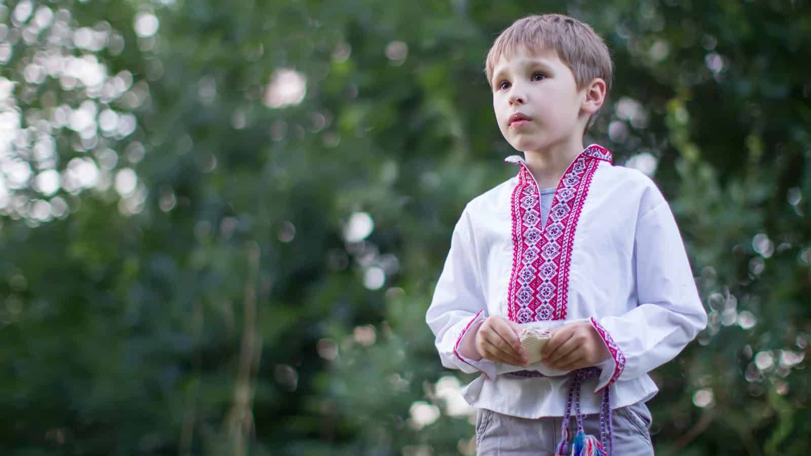 A young boy wearing a traditional embroidered white shirt stands outdoors, holding something in his hands, with green foliage in the background.