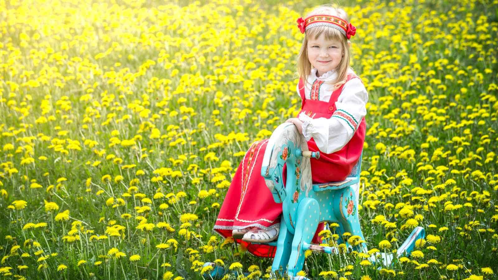 A young girl in traditional clothing sits on a painted rocking horse in a field of yellow dandelions.