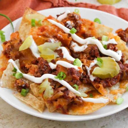 A plate of nachos topped with ground beef, melted cheese, pickle slices, sour cream, and chopped green onions, with tortilla chips and green onions beside the plate.