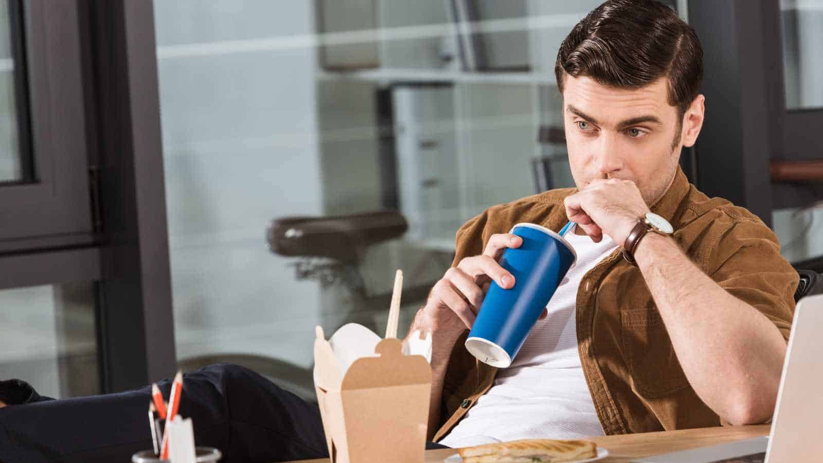 A man sits at a desk with his feet up, holding a blue cup and looking thoughtful. There are takeout food containers and a laptop on the desk.