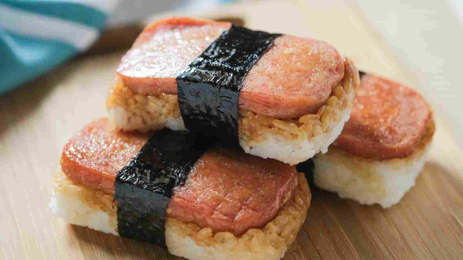 Three pieces of Spam musubi, each with a slice of Spam on rice, wrapped with a strip of seaweed, are arranged on a wooden surface.