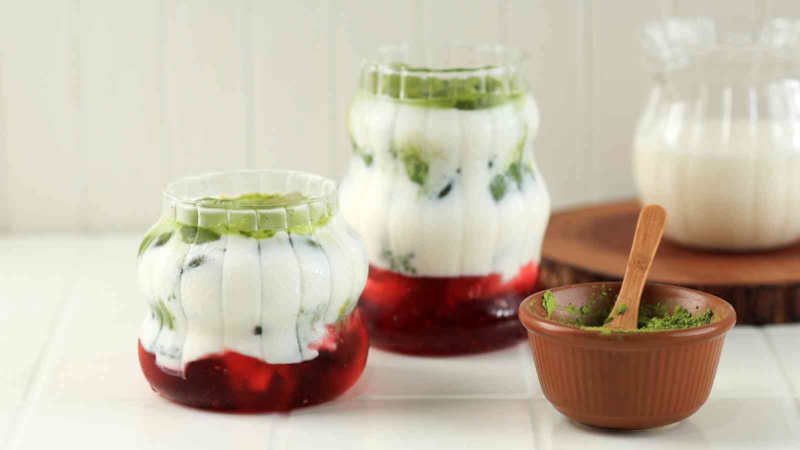 Two glasses filled with layers of red jelly, white yogurt, and green matcha, next to a small bowl of green powder with a spoon on a white surface.