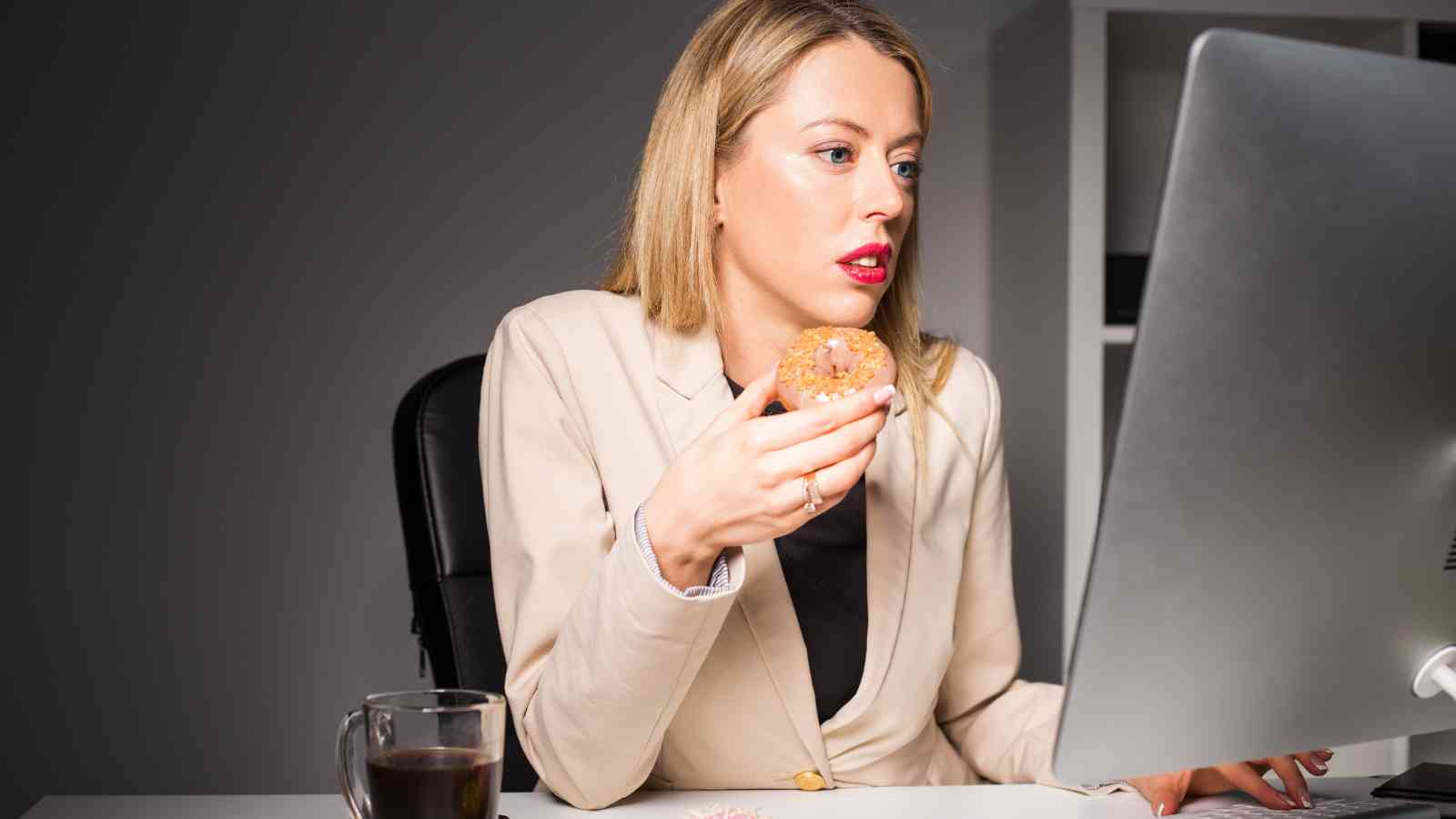 Woman in a beige suit sitting at a desk, eating a doughnut and looking intently at a computer monitor with a cup of coffee nearby.