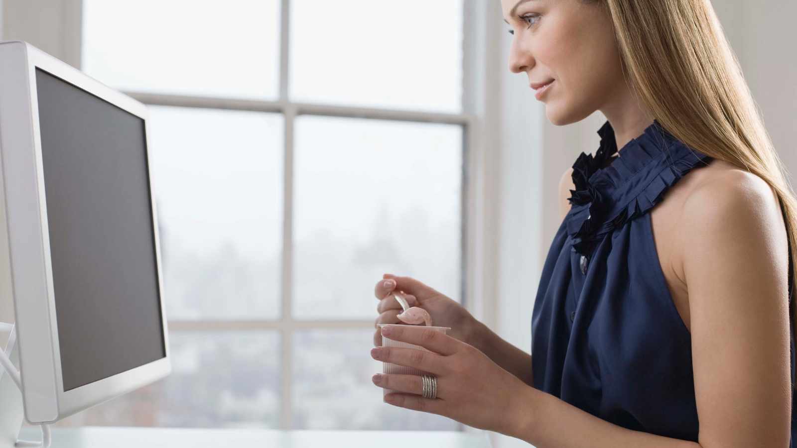 Woman holding a cup sits at a desk and looks at a computer monitor in a bright room with a large window in the background.