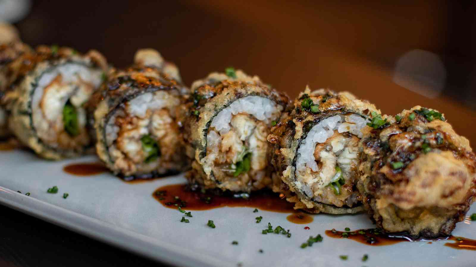Close-up of several pieces of deep-fried sushi roll topped with sauce and chopped herbs, arranged in a row on a rectangular plate.