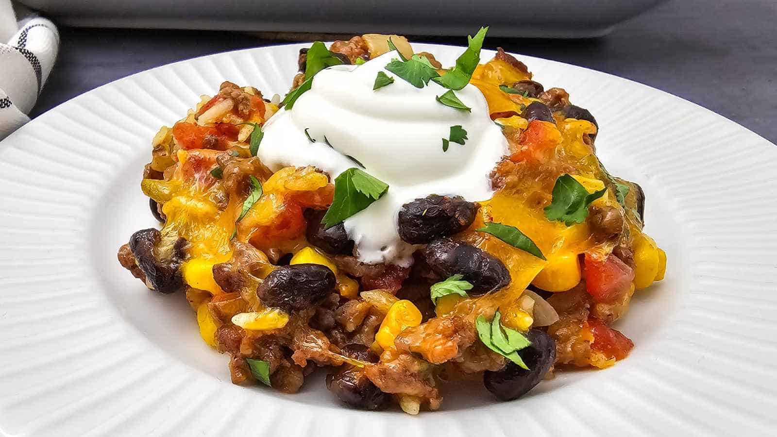 A serving of casserole with black beans, corn, and tomatoes, topped with a dollop of sour cream and chopped cilantro on a white plate.