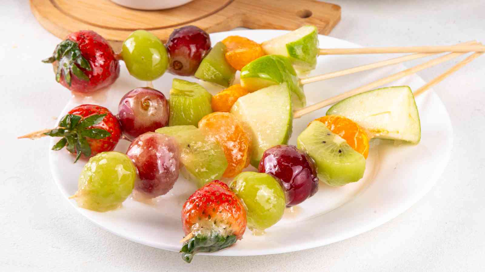 Several skewers of assorted candied fruit, including strawberries, grapes, kiwi, oranges, and apple slices, arranged on a white plate.