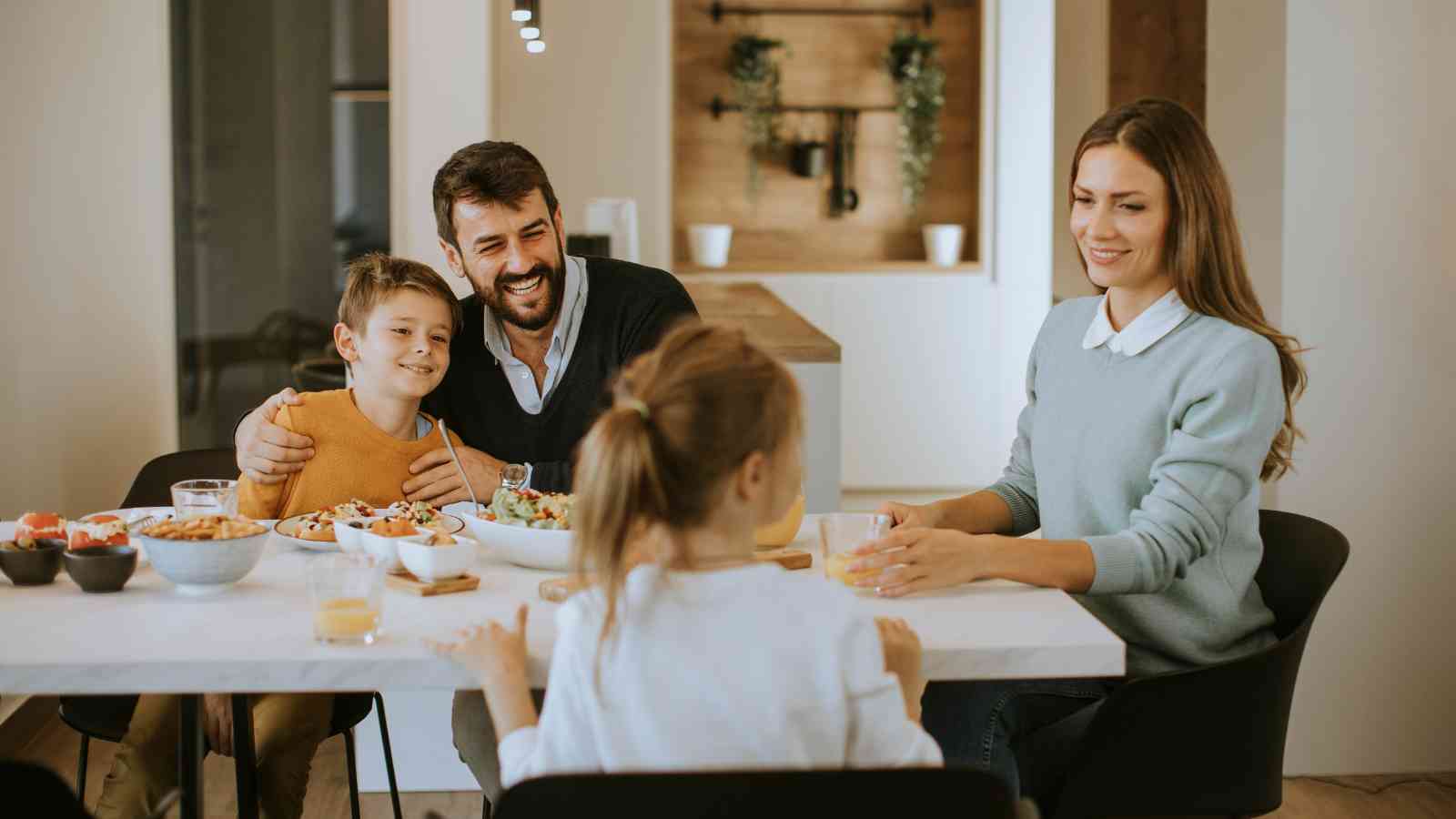 A family of four sits together at a dining table, sharing a meal in a modern, well-lit kitchen.