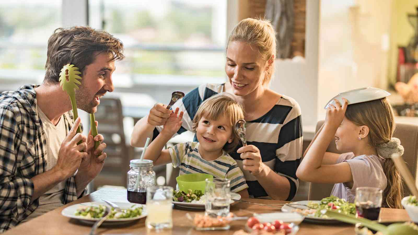A family of four sits at a dining table, smiling and playing with utensils and dishes while eating a meal together.