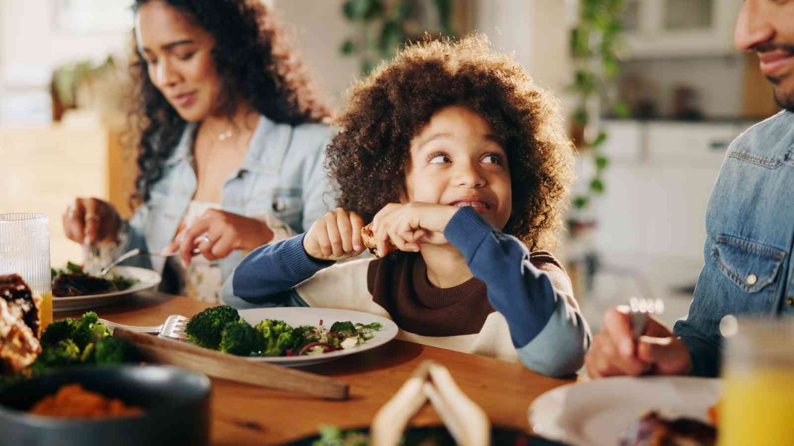 A young child smiles while eating a meal with two adults at a dining table, with plates of food and vegetables in front of them.