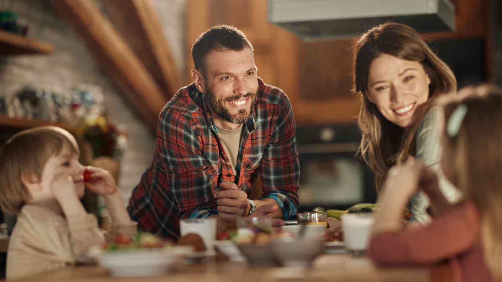 A family of four sits at a kitchen table, smiling and talking together while having a meal.